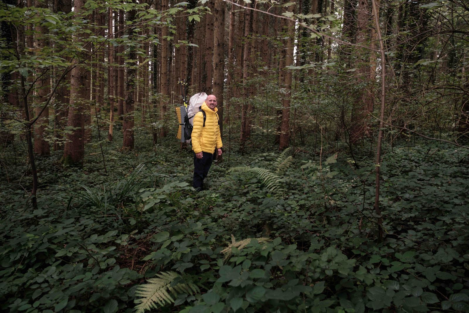 Erlebnispädagoge Manuel Seiler mit gelber Daunenjacke und Übernachtungsgepäck auf dem Rücken mitten im Dickicht im Wald